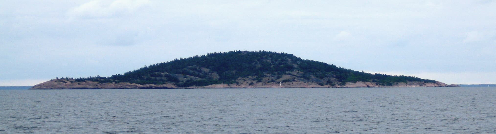 Den kupolformade granitön Blå Jungfrun sedd från havet, med klippig strandlinje och tät skog på toppen.