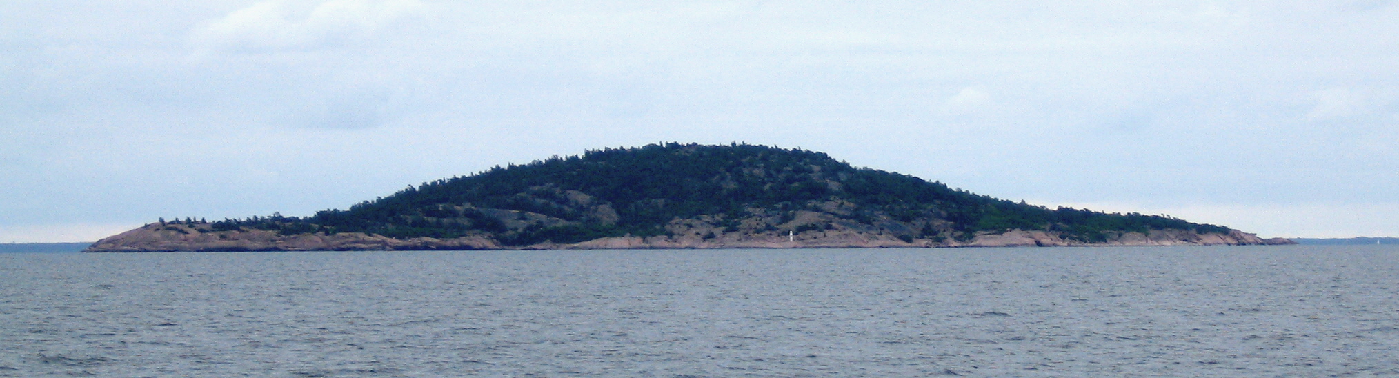 Den kupolformade granitön Blå Jungfrun sedd från havet, med klippig strandlinje och tät skog på toppen.